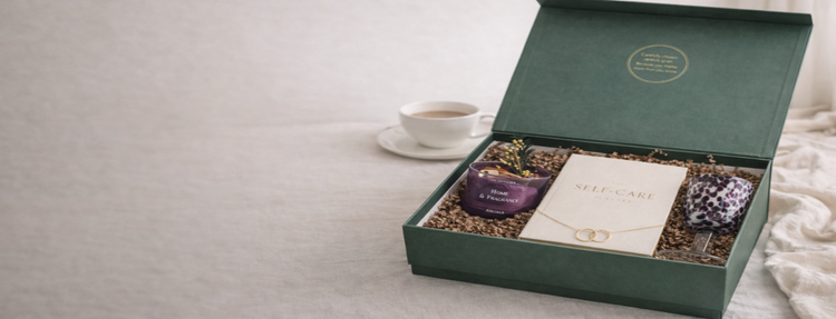 Green gift box with self-care items including a candle, necklace, and plant on a neutral background.